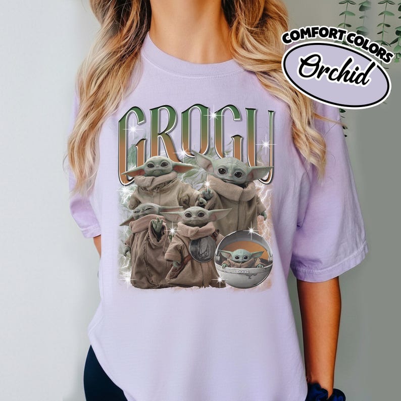 K&ouml;nnte beinhalten: Ein hellviolettes Kurzarm-T-Shirt mit dem Schriftzug GROGU in gro&szlig;en Buchstaben &uuml;ber einer Grafik von mehreren Grogu-Figuren in beigen Roben, darunter eine in einer schwebenden Kapsel, mit kleinen Sternakzenten und einem Comfort Colors Orchid-Etikett.