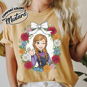 Peut inclure: T-shirt moutarde avec une illustration de dessin anim&eacute; d'un personnage aux cheveux tress&eacute;s, faisant un clin d'&oelig;il et saluant. Le personnage est entour&eacute; d'une couronne florale avec des roses rouges, des marguerites et des fleurs bleues. Le texte "COMFORT COLORS" et "Mustard" est en haut &agrave; gauche.