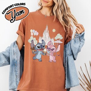 Puede incluir: Camiseta color &oacute;xido con una ilustraci&oacute;n de dibujos animados de Stitch y Angel sosteniendo conos de helado frente a un castillo. La camiseta tiene el texto "COMFORT COLORS" y "Yam" en un &oacute;valo marr&oacute;n.