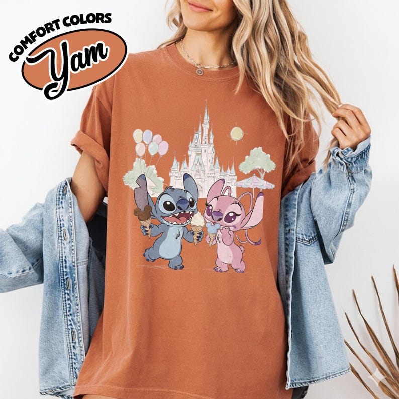 Puede incluir: Camiseta color &oacute;xido con una ilustraci&oacute;n de dibujos animados de Stitch y Angel sosteniendo conos de helado frente a un castillo. La camiseta tiene el texto "COMFORT COLORS" y "Yam" en un &oacute;valo marr&oacute;n.