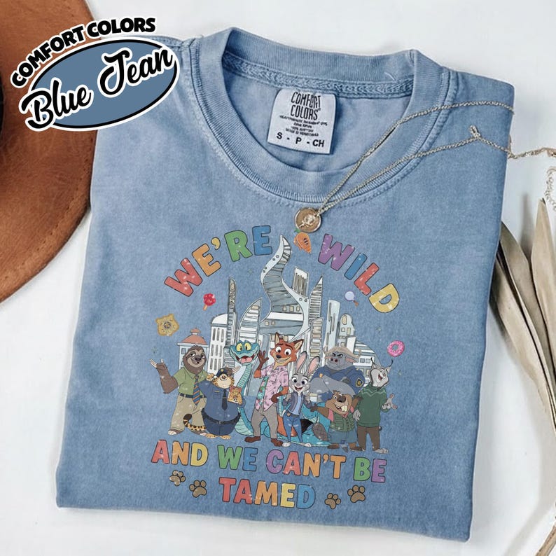 May include: A blue Comfort Colors t-shirt with the text "WE'RE WILD AND WE CAN'T BE TAMED" and a colorful graphic of cartoon characters. The shirt has a crew neck and short sleeves. A silver necklace is draped across the shirt.