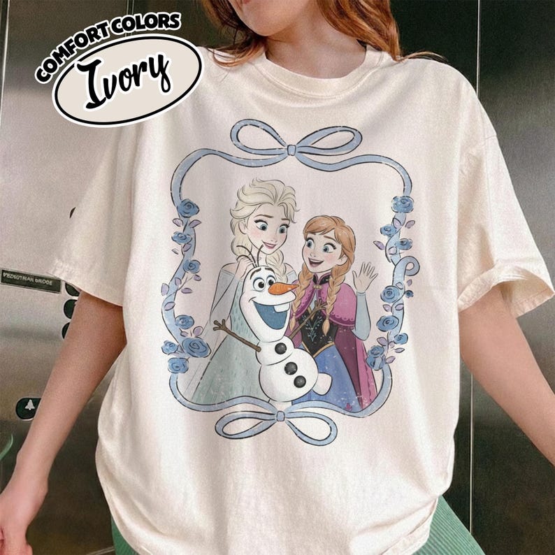 May include: Ivory-colored t-shirt featuring a graphic of Elsa, Anna, and Olaf from the movie Frozen, framed by a blue ribbon and floral design. The shirt has the words "Comfort Colors" and "Ivory" printed on the top left.