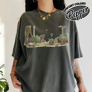 May include: Dark grey t-shirt featuring a cartoon scene with several Grogu characters in a forest setting. The shirt has the words "COMFORT COLORS" and "Pepper" printed on the upper right side. The shirt is a relaxed fit.