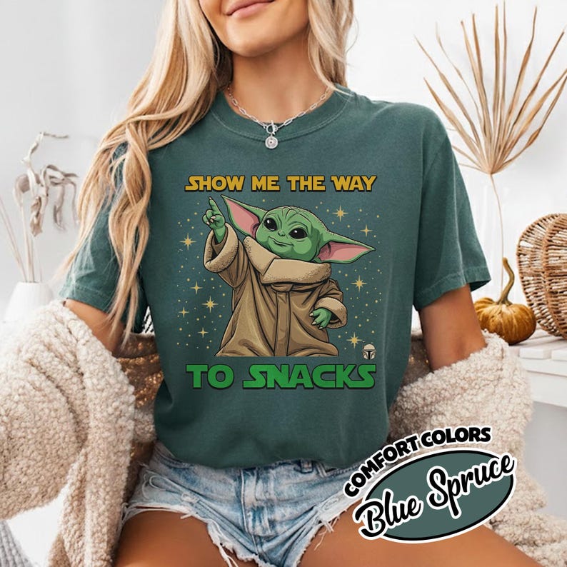 May include: A teal-colored t-shirt featuring a graphic of a green alien character with large ears, wearing a brown cloak. The text "SHOW ME THE WAY TO SNACKS" is above and below the character. The shirt also has the text "COMFORT COLORS Blue Spruce".