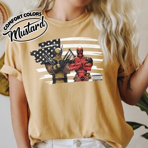 May include: A mustard yellow t-shirt featuring a graphic print of Wolverine and Deadpool standing in front of a stylized American flag. The text COMFORT COLORS MUSTARD is printed in the upper left corner, and a Parental Advisory label is visible.