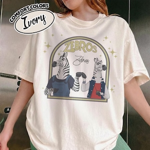 May include: Ivory t-shirt with a graphic of two zebras in a circular design, with the text "ZEBROS" and "Zebros". The zebras are wearing outfits. The shirt also has the text "COMFORT COLORS" and "Ivory".