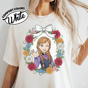 Peut inclure: T-shirt blanc cr&egrave;me avec un motif de couronne florale et un personnage de dessin anim&eacute; qui fait un clin d'&oelig;il et salue. La couronne comprend des roses rouges, des marguerites et des fleurs bleues, surmont&eacute;es d'un n&oelig;ud blanc. Le t-shirt porte les mots "Comfort Colors" et "White".