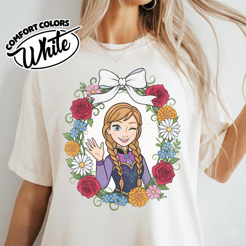 Peut inclure: T-shirt blanc cr&egrave;me avec un motif de couronne florale et un personnage de dessin anim&eacute; qui fait un clin d'&oelig;il et salue. La couronne comprend des roses rouges, des marguerites et des fleurs bleues, surmont&eacute;es d'un n&oelig;ud blanc. Le t-shirt porte les mots "Comfort Colors" et "White".
