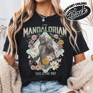 May include: A black t-shirt featuring a graphic print of The Mandalorian and Grogu surrounded by colorful flowers and butterflies. The text reads THE MANDALORIAN at the top and THIS IS THE WAY at the bottom. A Comfort Colors Black label is visible.