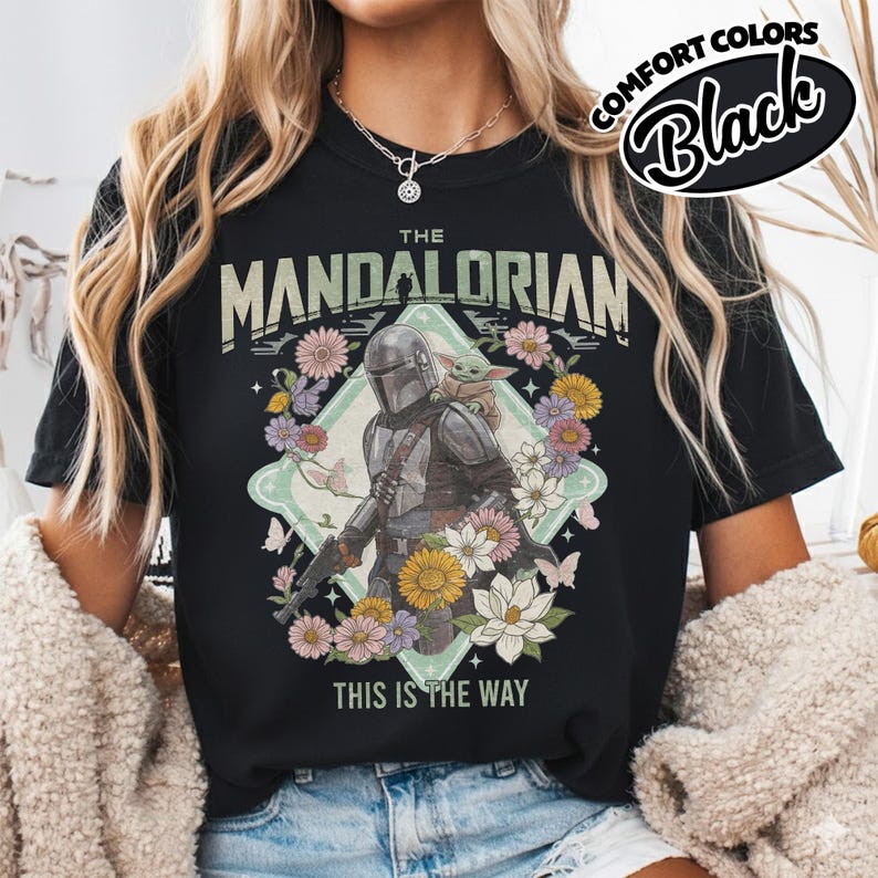 May include: A black t-shirt featuring a graphic print of The Mandalorian and Grogu surrounded by colorful flowers and butterflies. The text reads THE MANDALORIAN at the top and THIS IS THE WAY at the bottom. A Comfort Colors Black label is visible.