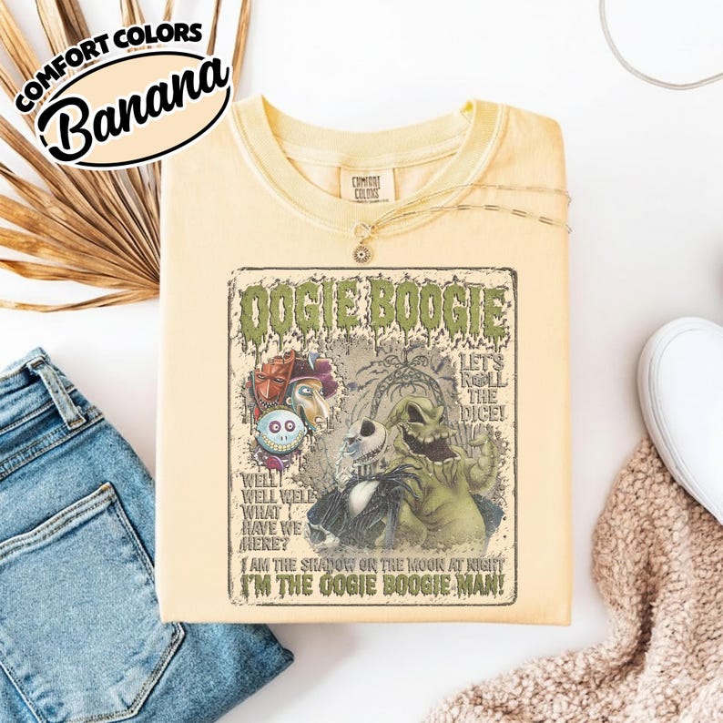 May include: A light yellow t-shirt with a graphic featuring characters and text from "The Nightmare Before Christmas." The graphic includes the words "Oogie Boogie" and "Let's Roll the Dice!" The shirt has a crew neck and is folded.