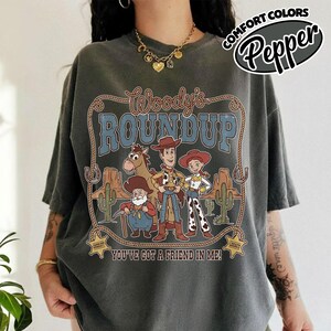 May include: Dark gray t-shirt featuring a graphic of Woody's Roundup characters in a desert scene. The design includes the text "Woody's Roundup" and "You've Got a Friend in Me!" in a Western-style font. The shirt has short sleeves.