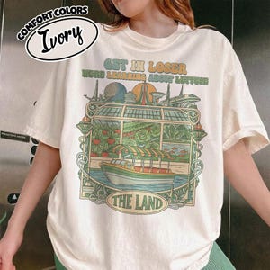 May include: Ivory-colored Comfort Colors t-shirt with a graphic featuring a boat, greenhouse, and text that reads "GET IN LOSER WE'RE LEARNING ABOUT LETTUCE" and "THE LAND". The shirt has a relaxed fit.