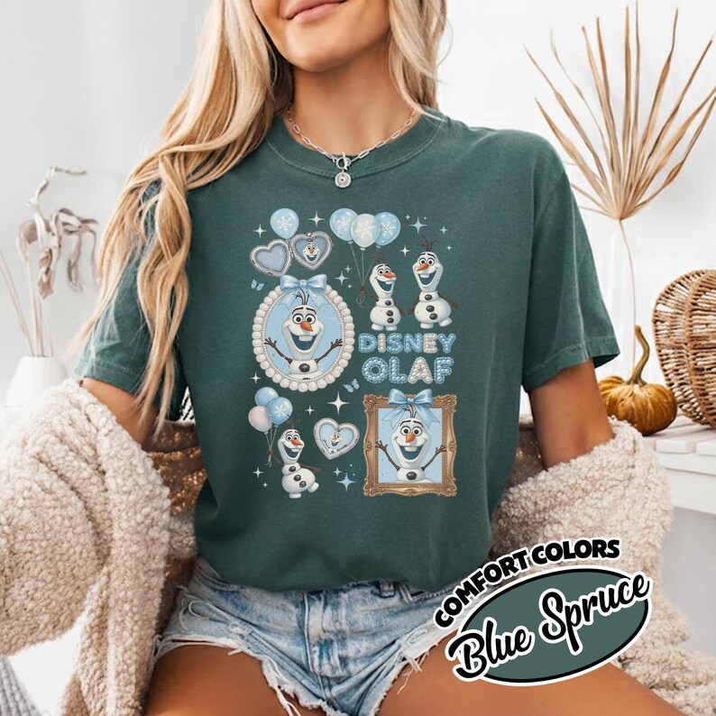 May include: A teal-colored t-shirt featuring a graphic of Olaf from Disney's Frozen. The design includes Olaf images, balloons, and the text "Disney Olaf." The shirt also has the text "COMFORT COLORS Blue Spruce" in a circular design.