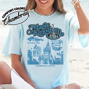 May include: Light blue t-shirt with a graphic of the Magic Kingdom. The design includes the words "Magic Kingdom" and illustrations of Autopia Car, Peter Pan's Flight Vehicle, Tiki Room Drummer, and Cinderella Castle.
