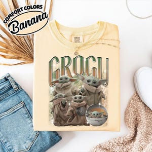 K&ouml;nnte beinhalten: Ein blassgelbes T-Shirt mit Rundhalsausschnitt und einem Grafikdruck der Figur Grogu in verschiedenen Posen. Der Text "GROGU" ist in gro&szlig;en, dunkelgr&uuml;nen Buchstaben &uuml;ber den Figuren gedruckt. Das Etikett zeigt "Comfort Colors" und "Banana".