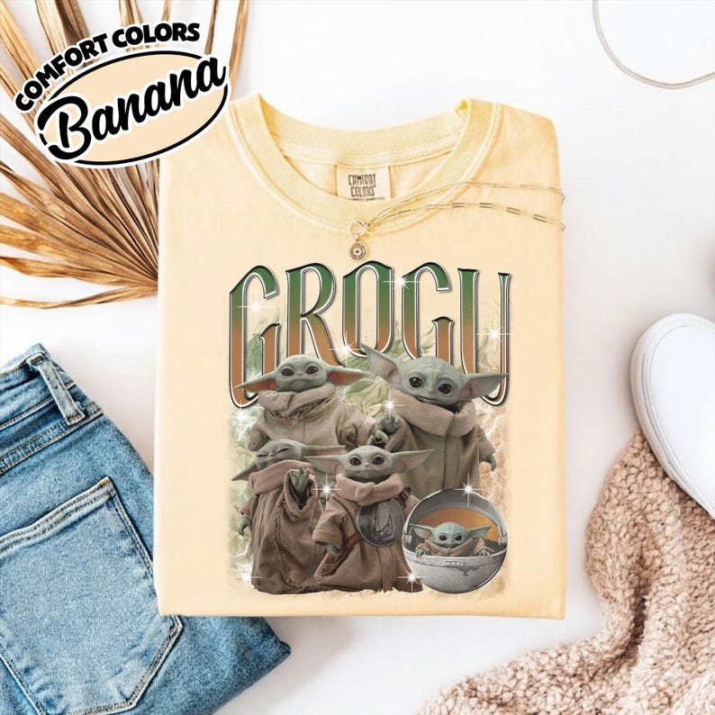 K&ouml;nnte beinhalten: Ein blassgelbes T-Shirt mit Rundhalsausschnitt und einem Grafikdruck der Figur Grogu in verschiedenen Posen. Der Text "GROGU" ist in gro&szlig;en, dunkelgr&uuml;nen Buchstaben &uuml;ber den Figuren gedruckt. Das Etikett zeigt "Comfort Colors" und "Banana".