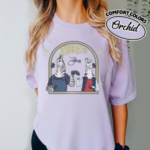 May include: Pale purple t-shirt with a cartoon illustration of two zebras inside a yellow arch, with the text "ZEBROS". The zebras are wearing dungarees and a jacket. The shirt also has the text "Comfort Colors" and "Orchid".