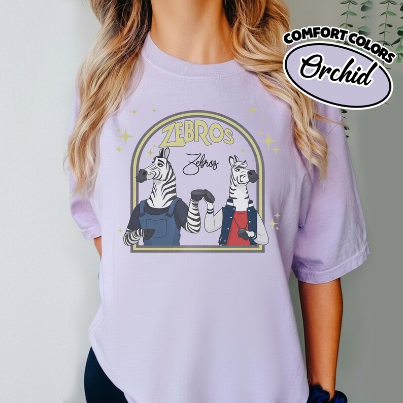 May include: Pale purple t-shirt with a cartoon illustration of two zebras inside a yellow arch, with the text "ZEBROS". The zebras are wearing dungarees and a jacket. The shirt also has the text "Comfort Colors" and "Orchid".