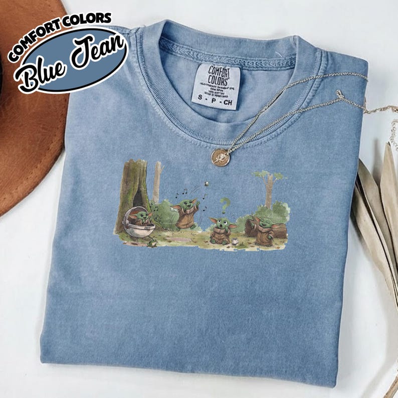 May include: Blue jean Comfort Colors t-shirt featuring a graphic of Baby Yoda in a forest scene. The shirt has a crew neck and short sleeves. The text "COMFORT COLORS" and "Blue Jean" are printed on the top left corner.