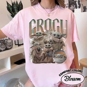 K&ouml;nnte beinhalten: Ein hellrosa Kurzarm-T-Shirt mit einem Grafikdruck von mehreren Grogu-Figuren in beigen Gew&auml;ndern. Der Text GROGU ist in einer gro&szlig;en, stilisierten gr&uuml;nen und orangefarbenen Schrift &uuml;ber den Figuren gedruckt. Eine Figur befindet sich in einer Kapsel.