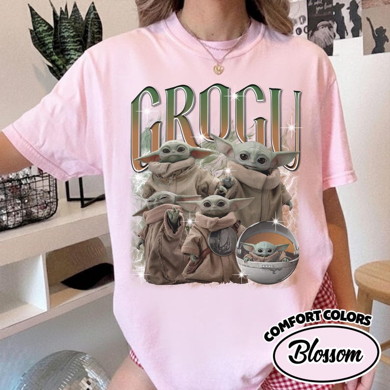 K&ouml;nnte beinhalten: Ein hellrosa Kurzarm-T-Shirt mit einem Grafikdruck von mehreren Grogu-Figuren in beigen Gew&auml;ndern. Der Text GROGU ist in einer gro&szlig;en, stilisierten gr&uuml;nen und orangefarbenen Schrift &uuml;ber den Figuren gedruckt. Eine Figur befindet sich in einer Kapsel.