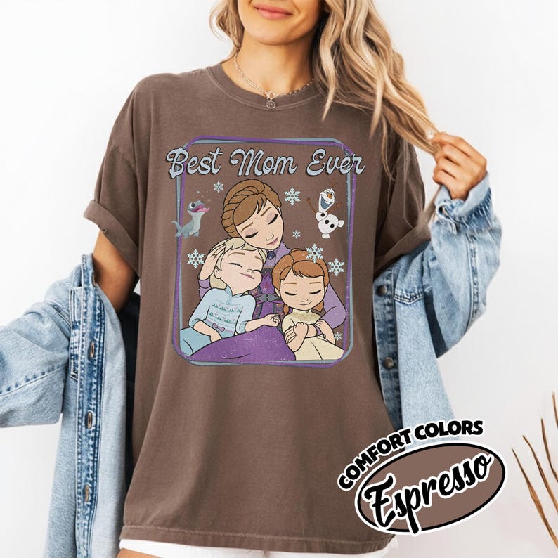 May include: Brown t-shirt with a graphic that says "Best Mom Ever" featuring an illustration of a mother and two children. The graphic includes snowflakes and characters. The shirt has short sleeves and a relaxed fit.