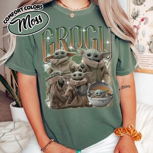 K&ouml;nnte beinhalten: Ein moosgr&uuml;nes T-Shirt mit dem Text COMFORT COLORS MOSS und GROGU in gro&szlig;er Schrift. Die Grafik zeigt vier kleine gr&uuml;ne Kreaturen mit gro&szlig;en Ohren und Augen, von denen eine in einer runden schwebenden Kapsel sitzt, umgeben von wei&szlig;en Blitzen.