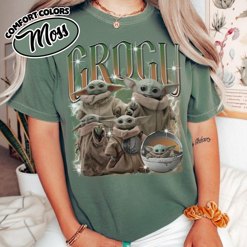 K&ouml;nnte beinhalten: Ein moosgr&uuml;nes T-Shirt mit dem Text COMFORT COLORS MOSS und GROGU in gro&szlig;er Schrift. Die Grafik zeigt vier kleine gr&uuml;ne Kreaturen mit gro&szlig;en Ohren und Augen, von denen eine in einer runden schwebenden Kapsel sitzt, umgeben von wei&szlig;en Blitzen.