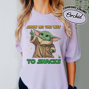 May include: Orchid Comfort Colors t-shirt featuring a graphic of a green alien character pointing upwards. The text reads "SHOW ME THE WAY TO SNACKS". The character is wearing a brown cloak. The shirt is light purple.