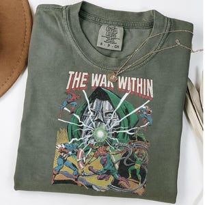 May include: Olive green t-shirt featuring a comic book graphic with the text "THE WAR WITHIN." The graphic depicts various comic book characters in action. A gold necklace with a pendant rests on the shirt.
