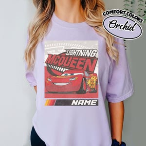 May include: A light purple t-shirt featuring a graphic of the red race car Lightning McQueen with the text Lightning McQueen and Name. The design includes a speedometer background and a yellow, orange, and red stripe accent at the bottom.