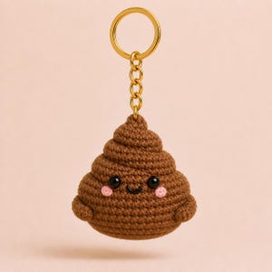 May include: A brown crocheted poop emoji keychain with a gold-colored keyring and chain. The emoji has a smiling face with black eyes and pink cheeks. The keychain is a fun and whimsical accessory.