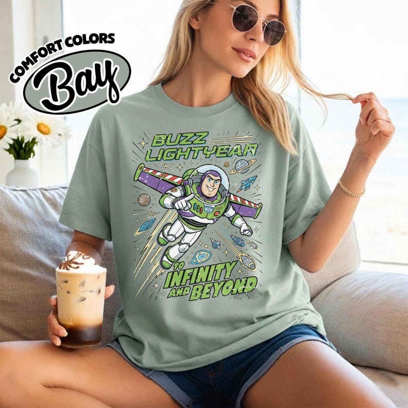 May include: A sage green t-shirt featuring a graphic of Buzz Lightyear flying through space with the text "BUZZ LIGHTYEAR TO INFINITY AND BEYOND". The design includes planets, stars, and a rocket ship in shades of purple, green, and blue.
