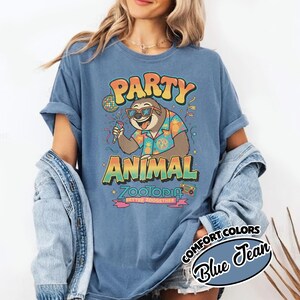 Puede incluir: Camiseta azul con un dise&ntilde;o de un perezoso con gafas de sol y una camisa hawaiana, sosteniendo un micr&oacute;fono. El texto dice "PARTY ANIMAL Zootopia" en letras retro coloridas. La camiseta est&aacute; etiquetada como "Comfort Colors Blue Jean".