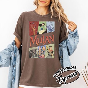 Puede incluir: Camiseta marr&oacute;n con un gr&aacute;fico de Mulan al estilo c&oacute;mic. La palabra "Mulan" es naranja. La camiseta tiene mangas enrolladas y una etiqueta Comfort Colors. La camiseta es de color marr&oacute;n oscuro.