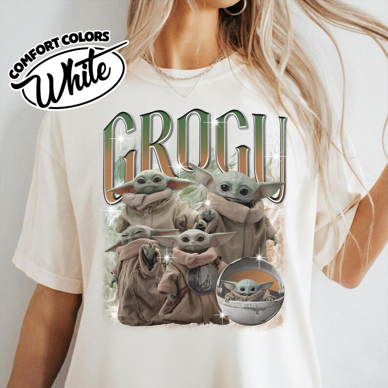 K&ouml;nnte beinhalten: Ein wei&szlig;es T-Shirt mit einem Grafikdruck der Figur Grogu in verschiedenen Posen. Der Text GROGU ist in gro&szlig;en, stilisierten Buchstaben &uuml;ber den Figuren gedruckt. Das Design enth&auml;lt kleine sternf&ouml;rmige Glanzeffekte und ein rundes Kapsel-Element.
