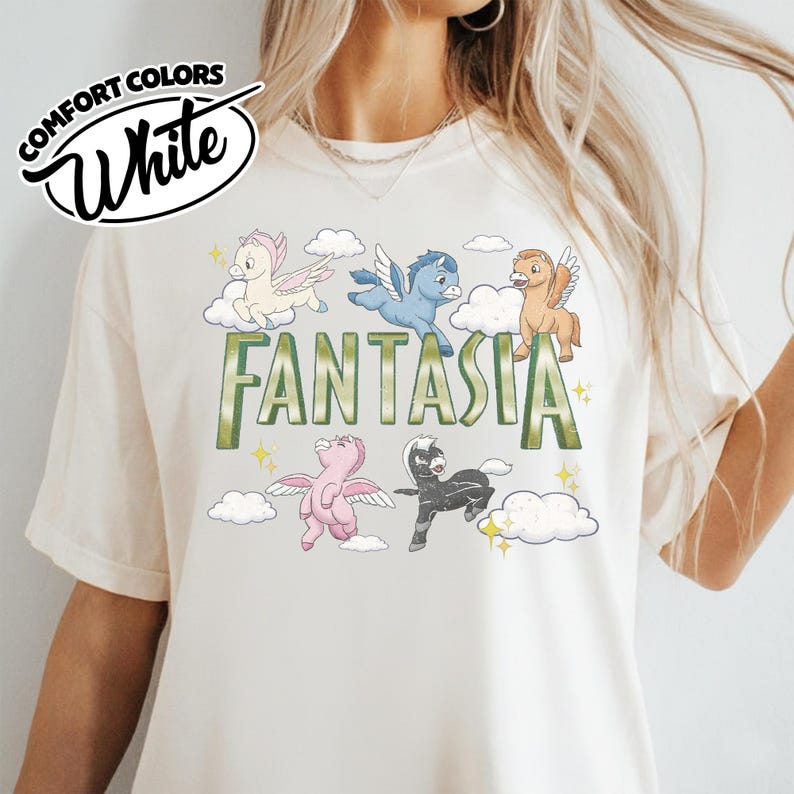 May include: Cream-coloured Comfort Colors t-shirt with the word "Fantasia" in green, and cartoon pegasus characters. The shirt has a relaxed fit and a crew neck.