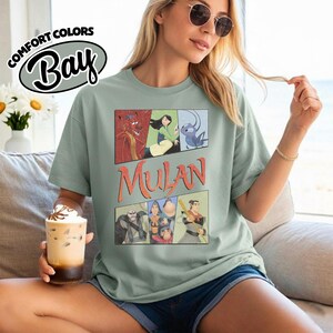 Puede incluir: Camiseta verde claro con un gr&aacute;fico de personajes de Mulan al estilo c&oacute;mic. La palabra "Mulan" est&aacute; impresa en naranja. La camiseta tambi&eacute;n tiene el texto "Comfort Colors Bay" en un &oacute;valo blanco.