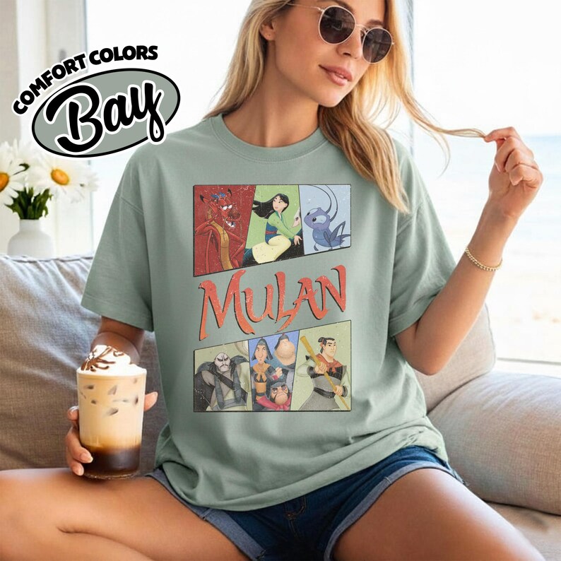 Puede incluir: Camiseta verde claro con un gr&aacute;fico de personajes de Mulan al estilo c&oacute;mic. La palabra "Mulan" est&aacute; impresa en naranja. La camiseta tambi&eacute;n tiene el texto "Comfort Colors Bay" en un &oacute;valo blanco.