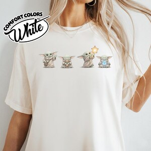 以下が含まれることがあります： 白いComfort ColorsのTシャツ。4つのGroguのイラストが描かれています。イラストは、バッグ、テディベア、星、光るオブジェクトを持ったGroguを描いています。シャツには「Comfort Colors」と「White」の文字が円形にデザインされています。
