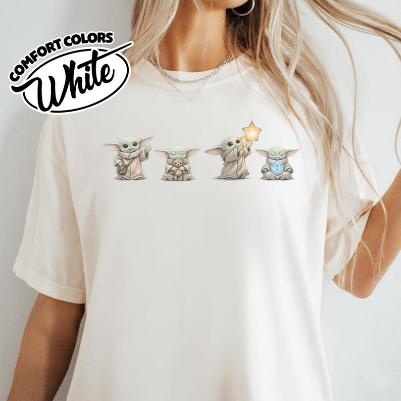 以下が含まれることがあります： 白いComfort ColorsのTシャツ。4つのGroguのイラストが描かれています。イラストは、バッグ、テディベア、星、光るオブジェクトを持ったGroguを描いています。シャツには「Comfort Colors」と「White」の文字が円形にデザインされています。