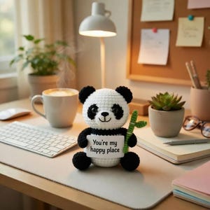 May include: A crocheted panda bear with black and white fur holds a sign that reads "You're my happy place." The panda is sitting on a desk with a cup of coffee, a keyboard, and a succulent plant in the background.