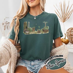 May include: A teal-colored t-shirt featuring a graphic of a character from a popular science fiction series. The graphic depicts the character in a forest setting with trees and a question mark. The text "COMFORT COLORS Blue Spruce" is also visible.