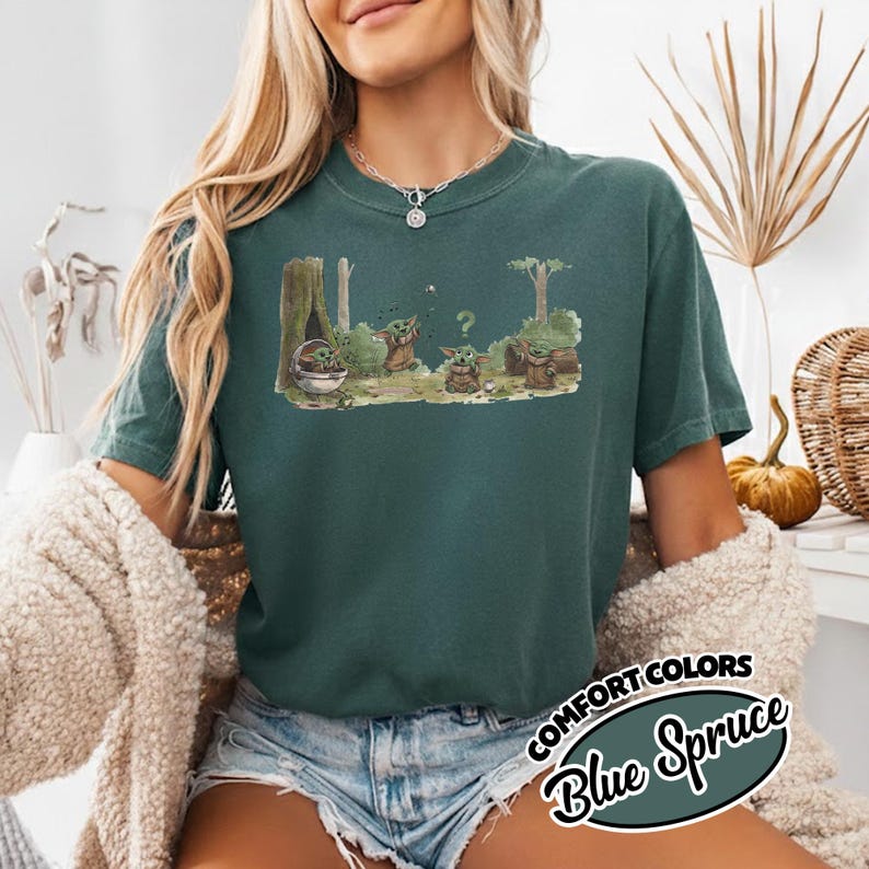 May include: A teal-colored t-shirt featuring a graphic of a character from a popular science fiction series. The graphic depicts the character in a forest setting with trees and a question mark. The text "COMFORT COLORS Blue Spruce" is also visible.