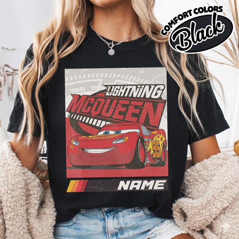 May include: A black t-shirt featuring a graphic print of the red race car Lightning McQueen. The design includes the text LIGHTNING MCQUEEN in bold red letters, a speedometer background, and a bottom bar with the word NAME and a tri-color stripe.