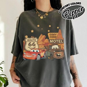 May include: Dark gray t-shirt featuring a graphic of Lightning McQueen and Mater from the movie Cars, with a Route 66 sign and Cozy Cone Motel. The shirt has the words "Comfort Colors" and "Pepper" printed on it.