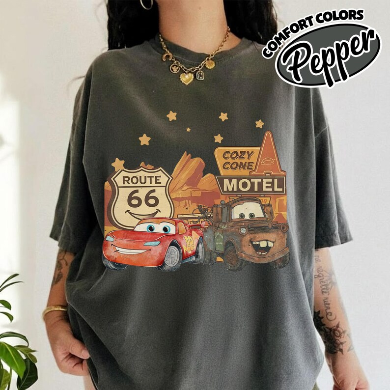 May include: Dark gray t-shirt featuring a graphic of Lightning McQueen and Mater from the movie Cars, with a Route 66 sign and Cozy Cone Motel. The shirt has the words "Comfort Colors" and "Pepper" printed on it.