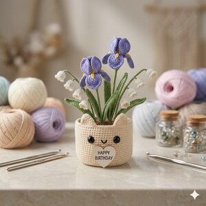 May include: A crocheted cat-shaped planter with purple iris and white lily of the valley flowers. The planter has a heart-shaped tag that reads "HAPPY BIRTHDAY". Crochet hooks and yarn balls in various colors are in the background.
