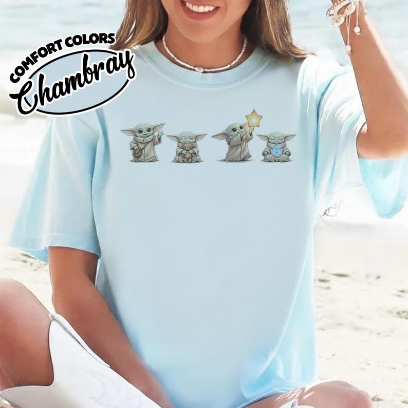 以下が含まれることがあります： 「COMFORT COLORS Chambray」の文字と4体のグローグーのキャラクターが描かれた水色のTシャツ。キャラクターは様々なポーズをしており、そのうちの1体は星を持っています。Tシャツは半袖でゆったりとしたフィット感です。